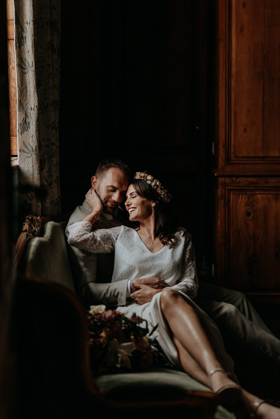 Mariés enlacés sur un canapé, la mariée rit, lumière de fenêtre