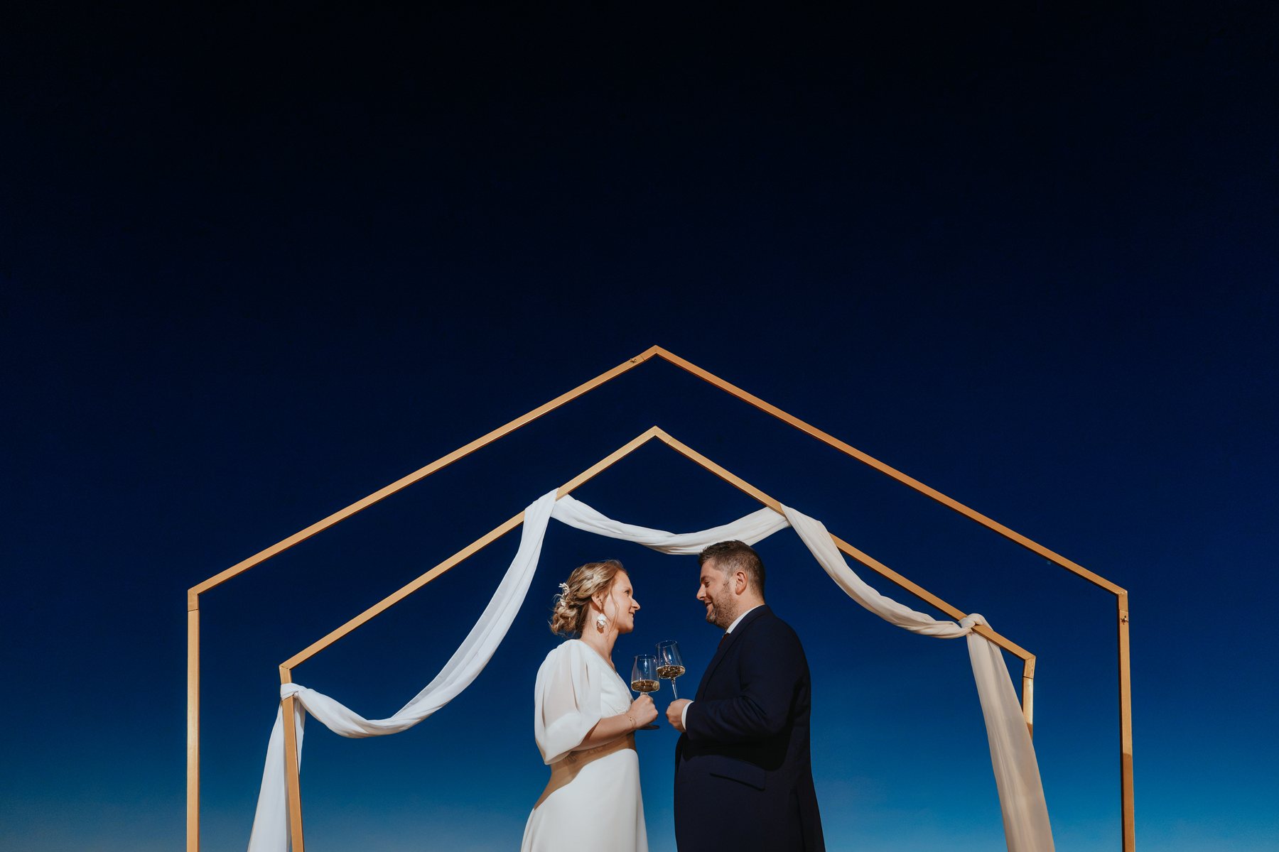 Clothilde et Erwan trinquent sous une arche dorée, ciel bleu
